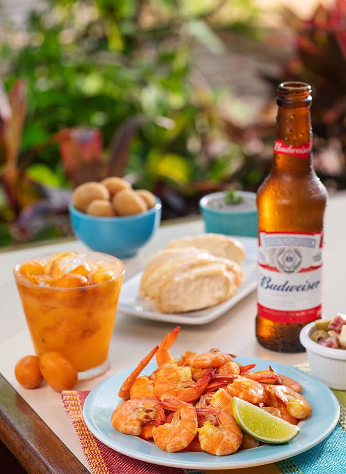 Em um dos ambientes do Salinas Maceió, está uma mesa com comidas e bebidas oferecidas no sistema all inclusive. Nela contém uma cerveja long neck, drinks e porções de petiscos.